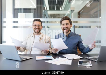 Due partner commerciali maschili che celebrano un progetto di successo, sorridono e tengono documenti durante una moderna riunione d'ufficio, trasmettendo lavoro di squadra, risultati e collaborazione professionale Foto Stock