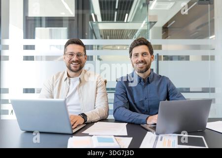 Due colleghi maschi sorridenti collaborano a una scrivania di coworking luminosa, i laptop si aprono e i documenti si diffondono durante la pianificazione di un progetto di avvio, discutendo strategie e idee Foto Stock