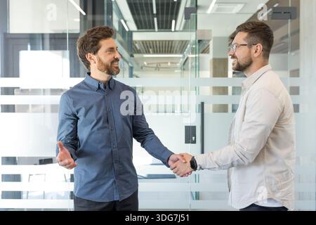 Due imprenditori maschi che stringono la mano e sorridono, mostrando un accordo commerciale di successo, una partnership e un saluto professionale in un moderno ambiente aziendale Foto Stock