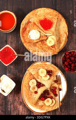 Deliziosi pancake russi con formaggio morbido, banane, caviale rosso e mirtilli rossi sul tavolo della cucina. Una sana colazione preparata con ingredienti freschi Foto Stock