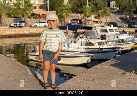 Ragazzo sicuro con cappello e camicia a righe in piedi sul molo del porticciolo mediterraneo con barche e palme nelle soleggiate giornate estive. Concetto di po Foto Stock