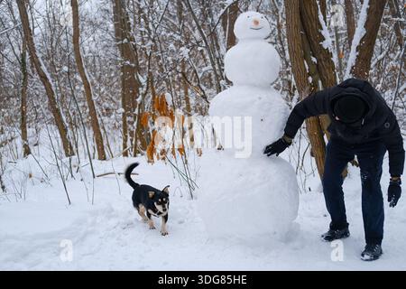 Vista orizzontale di un alto pupazzo di neve a tre livelli nella foresta innevata, cane che cammina sulla neve compatta a sinistra, persona con abiti scuri che leviga la superficie della neve. Foto Stock