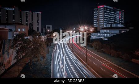 Una scena urbana notturna catturata con una lunga esposizione, che mostra percorsi luminosi fluenti dal traffico lungo una strada curva, incorniciata da moderni edifici per uffici Foto Stock