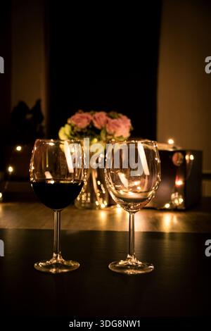 Due bicchieri da vino su un tavolo, uno pieno di vino rosso, calde luci bokeh e fiori sullo sfondo per un'atmosfera romantica. Foto Stock
