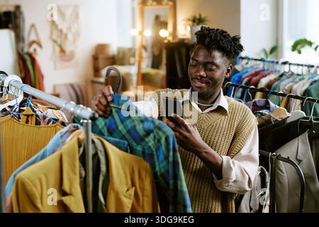 Giovane adulto, uomo nero che controlla l'abbigliamento di seconda mano mentre utilizza lo smartphone Foto Stock