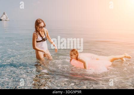 Madre figlia vacanza famiglia giocare con l'anello rosa nuoto nelle acque cristalline del mare su una spiaggia di ciottoli durante la soleggiata giornata estiva Foto Stock