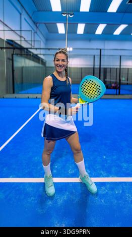 Un giocatore di padel che indossa una gonna in padel e la parte superiore posa felicemente, sorridente, in piedi su un campo blu con una racchetta. Foto Stock
