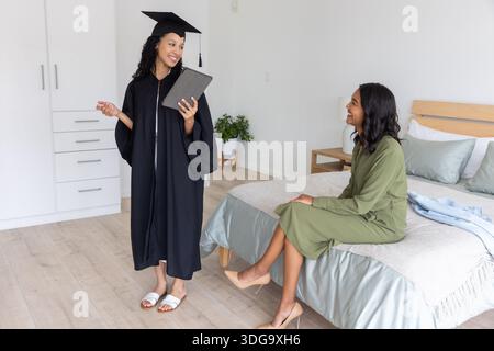 Mamma e figlia adolescente diverse in piedi in camera da letto con un tablet mentre indossano un abito da laurea Foto Stock