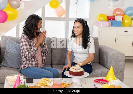 Mamma e figlia adolescente diverse applaudono e tengono una torta di compleanno sul divano del soggiorno Foto Stock