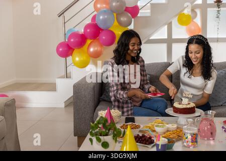 Mamma e figlia adolescente diverse che affettano la torta con la candela nel soggiorno in mezzo a palloncini Foto Stock