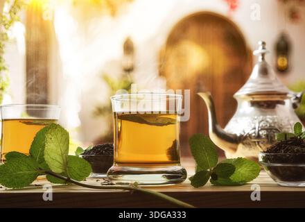 Dettagli di versare il tè marocchino da una teiera metallica a un bicchiere su un tavolo di legno con ciotola di tè a foglia secca decorata con foglie di menta secondo una tradizione Foto Stock