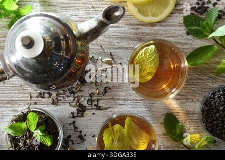 Dettaglio del tè verde in tazze di vetro su un tavolo da cucina in legno con ciotole contenenti foglie di tè essiccate, rametti di menta fresca e una teiera in metallo decorata Foto Stock
