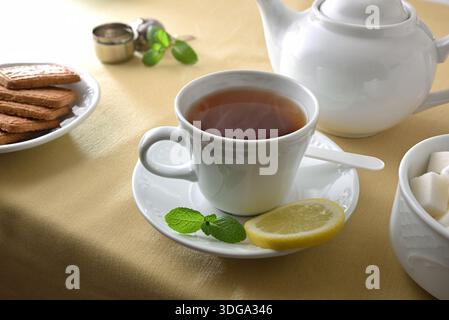 Tè verde in una tazza di ceramica bianca su un piatto con menta e limone su un tavolo con tovaglia marrone con contenitori e biscotti. Vista rialzata. Foto Stock