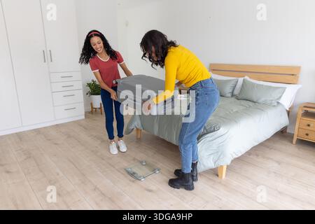 Mamma e figlia adolescente diverse che sollevano la valigia grigia in piedi su una bilancia digitale in camera da letto Foto Stock