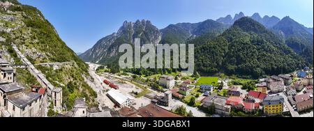 Vista aerea delle aspre montagne incontra una pittoresca cittadina con edifici dal tetto rosso e un antico complesso minerario, Cave del Predil, Friuli-Venezia Giulia, Italia. Foto Stock
