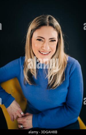 Donna sorridente seduta su una sedia gialla che guarda la macchina fotografica Foto Stock