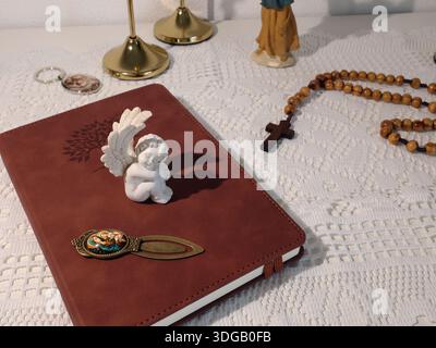 Angolo cattolico a casa. Figurina d'angelo, perline di rosario in legno, croce e libro di preghiere. Fede, simboli e tradizioni. Foto Stock
