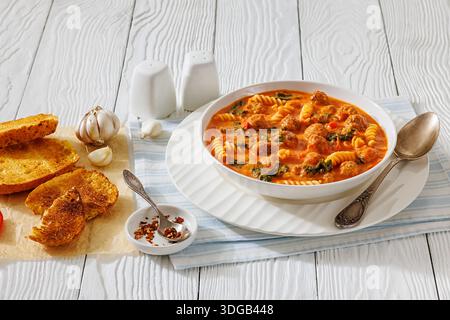 Zuppa italiana di pasta fusilli con polpette e spinaci in crema salsa di pomodoro in ciotola bianca su tavola di legno bianco con pane tostato e cucchiaio, orizzonte Foto Stock
