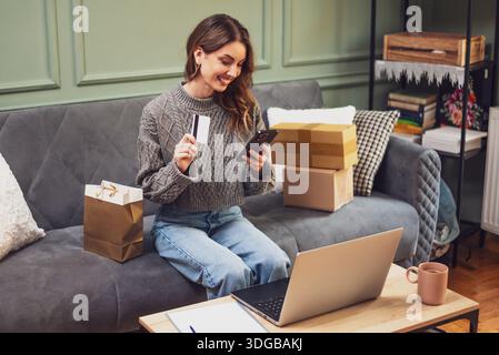 Comodamente seduto su un divano, la donna mostra la sua carta di credito mentre usa il suo smartphone per lo shopping online. Foto Stock