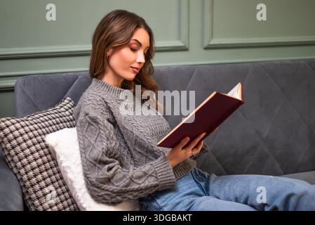 Una donna adulta rilassata si siede sul divano a casa e legge un libro. Foto Stock