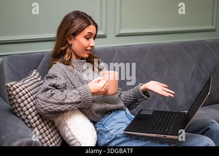 Una donna pensiva si rilassa su un divano con una bevanda calda in mano, lavorando sul suo computer portatile. Foto Stock