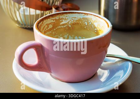 Tazza da caffè rosa vuota con residui di latte sul piattino con cucchiaio e decorazioni sfocate sul tavolino da caffè illuminato dal sole Foto Stock