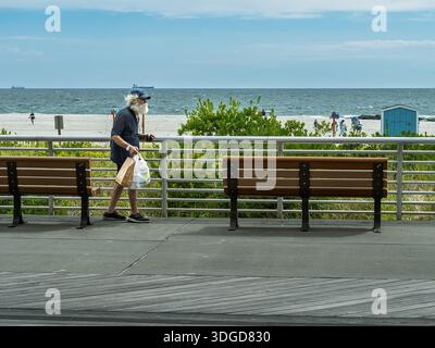 Un uomo anziano cammina lungo la passerella di Long Beach, New York, portando con sé borse della spesa mentre l'attività in spiaggia continua sullo sfondo. Foto Stock