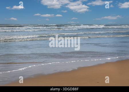 Una tranquilla scena costiera caratterizzata da dolci onde marine che si infrangono su una spiaggia di sabbia liscia sotto un cielo blu Foto Stock