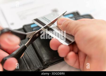 Primo piano di una persona che usa un paio di forbici per tagliare una carta di credito con una borsa e banconote della carta di credito sullo sfondo. Foto Stock