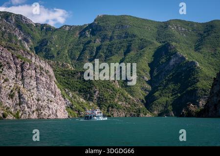 Un traghetto passeggeri naviga attraverso le acque turchesi del lago Komani, circondato da ripide montagne coperte da foreste e scogliere rocciose Foto Stock