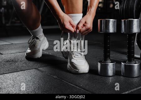 Primo piano di una donna che lega lacci delle scarpe in palestra prima di allenarsi, preparazione motivante al fitness con manubri e attrezzatura da allenamento Foto Stock