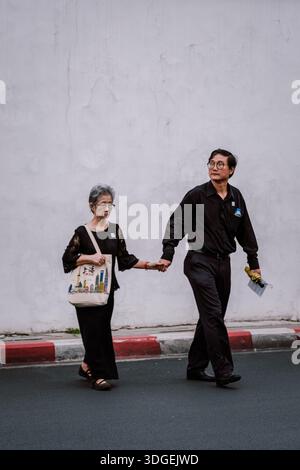 Bangkok, Thailandia, 19 novembre 2025. Una coppia di anziani si diverte a passeggiare insieme, tenendosi per mano mentre esplora le strade animate, circondate da Foto Stock