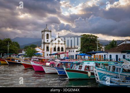 Barche colorate ormeggiate nel porto di Paraty con Igreja de Santa Rita de Cássia sotto cieli drammatici - Paraty, Rio de Janeiro, Brasile Foto Stock