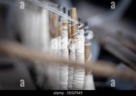 Fila di bobine di filo bianco avvolte su mandrini in legno, pronte per l'uso nella produzione tessile, evidenziando l'artigianalità tradizionale e la produzione Foto Stock