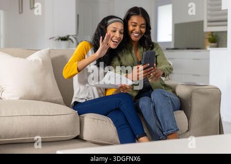 Mamma e figlia adolescente diverse che partecipano a una chiamata virtuale sul divano che regge lo smartphone Foto Stock