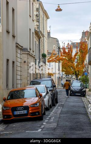 Bordeaux, Francia - 23 novembre 2021: Una strada stretta di Bordeaux presenta auto parcheggiate, tra cui un'Audi arancione, un uomo con una giacca arancione che cammina, traditore Foto Stock