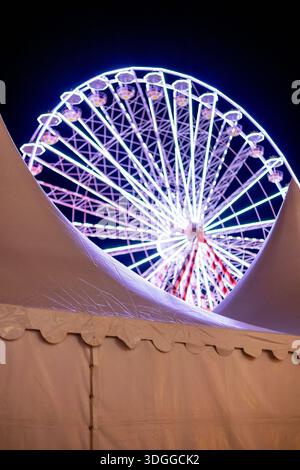 Vista notturna astratta verticale di una ruota panoramica illuminata con luci al neon sfocate dietro la cima di una grande tenda bianca in una fiera della città a W Foto Stock