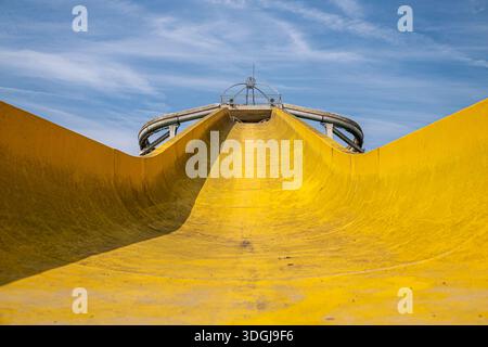 Prospettiva ad angolo elevato guardando verso il basso la curva di uno scivolo d'acqua giallo asciutto e sporco. L'immagine cattura il vuoto e la desolazione di una riproduzione defunta Foto Stock