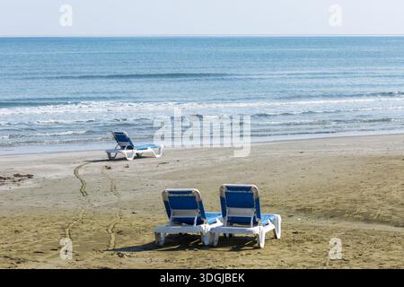 Numerosi lettini da spiaggia bianchi e blu vuoti riposano su un'ampia distesa di spiaggia sabbiosa. I lettini vuoti simboleggiano un invito pacifico al relax a. Foto Stock
