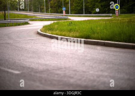 Una visuale ad angolo basso di una rampa curva per asfalto su uno svincolo autostradale. La strada è delimitata da un marciapiede di cemento e da un verde verde verde con i cartelli stradali i. Foto Stock