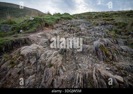Un ampio angolo di un aspro sentiero escursionistico coperto di pietra che sale su una collina erbosa sotto un cielo coperto sul Ben Lomond in Scozia. Foto Stock