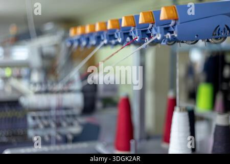 Vista ravvicinata e selettiva della guida filettata su una macchina per maglieria industriale. I fili rossi e bianchi vengono alimentati attraverso i sensori dell'au Foto Stock