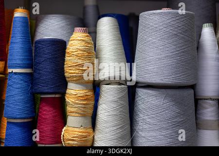 Una disposizione colorata e ravvicinata di coni in filato in una fabbrica di maglieria. Grandi bobine di filetto blu, giallo, grigio e rosso sono impilate su un ripiano, mostrare Foto Stock