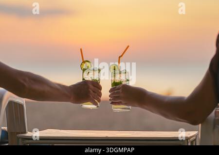 Le mani degli amanti tengono i bicchieri da cocktail mojito sullo sfondo di un tramonto sul mare. Festeggiano San Valentino in vacanza al mare o in luna di miele e.. Foto Stock