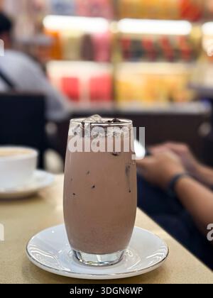 Alto bicchiere di cioccolato ghiacciato ricoperto di trucioli in un'accogliente caffetteria con sfondo sfocato e tazza da caffè. Foto Stock