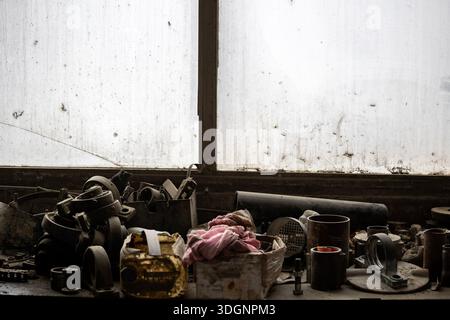 Banco da lavoro ingombrante prima di una finestra sporca con parti metalliche: Ingranaggi, tubi, raccordi. Attrezzi manuali sparsi e un panno rosa deformato in un cartone usurato Foto Stock