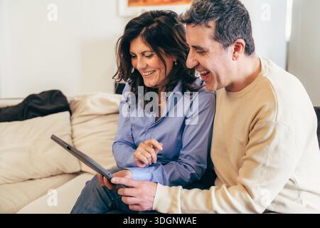 Felice coppia di mezza età seduta vicino su un divano, sorridente e godendo un momento di relax insieme a casa. Una calda scena di stile di vita sulla connessione, Happi Foto Stock