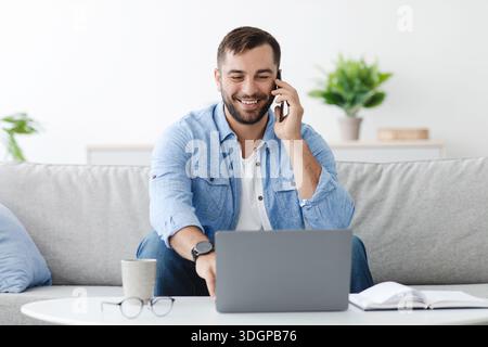 Un uomo europeo sorridente e maturo con la barba che parla al telefono, guarda il laptop nell'interno del salotto Foto Stock
