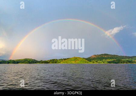 Un vivace arcobaleno si affaccia sul lago Mutanda a Kisoro, nel sud-ovest dell'Uganda, mentre la luce del sole si irrompe attraverso le nuvole di pioggia e si riflette sull'altopiano calmo Foto Stock