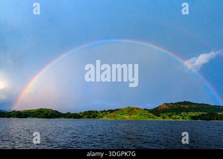 Un vivace arcobaleno si affaccia sul lago Mutanda a Kisoro, nel sud-ovest dell'Uganda, mentre la luce del sole si irrompe attraverso le nuvole di pioggia e si riflette sull'altopiano calmo Foto Stock